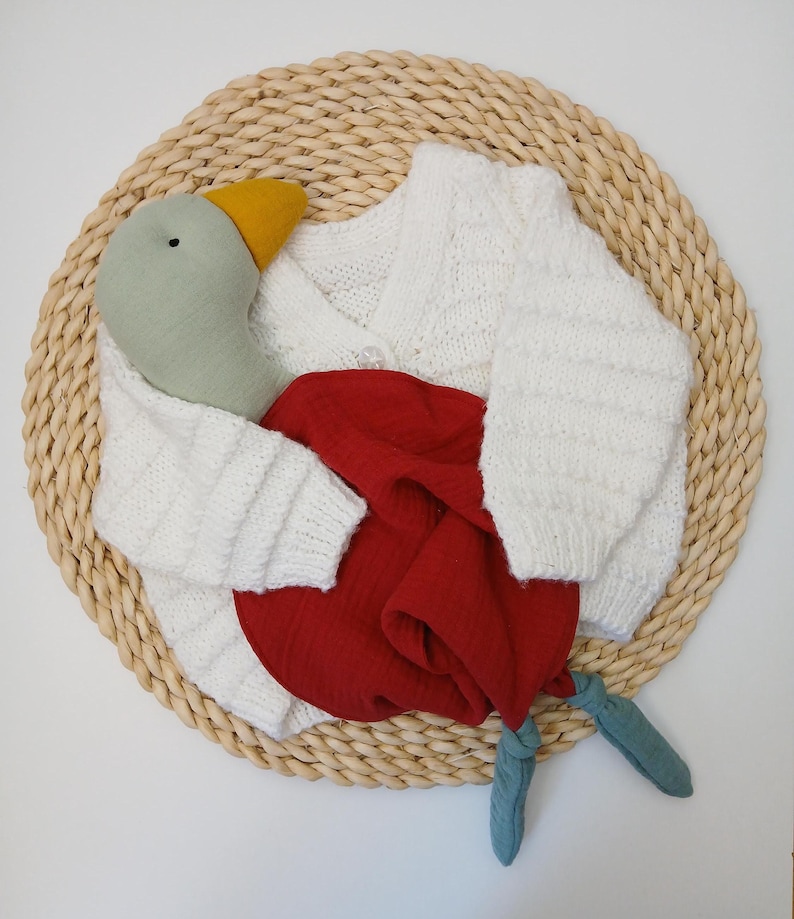 May include: A white knitted cardigan with a button closure, paired with a red fabric and a stuffed goose toy. The goose has a green head, yellow beak, and blue feet. The items are arranged on a woven, circular mat.