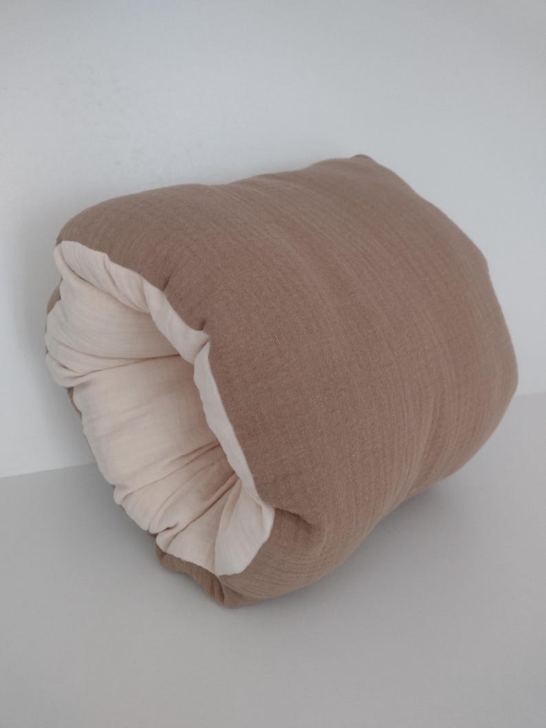 May include: A soft, rectangular cushion featuring a textured, taupe-coloured exterior fabric and a contrasting cream-coloured interior lining. The cushion is folded over itself, creating a rounded, tube-like shape that reveals both colour layers.