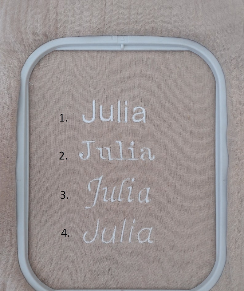 K&ouml;nnte beinhalten: Nahaufnahme eines hellbeigen Stoffes mit dem Namen "Julia" in Wei&szlig; in vier verschiedenen Schriftarten gestickt. Der Stoff wird in einem rechteckigen Stickrahmen gehalten.