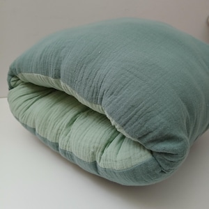May include: A soft, square-shaped cushion featuring a dual-tone design with a sage green exterior and a lighter mint green interior. The fabric has a visible crinkled, textured finish characteristic of muslin, and the cushion is folded slightly inward.