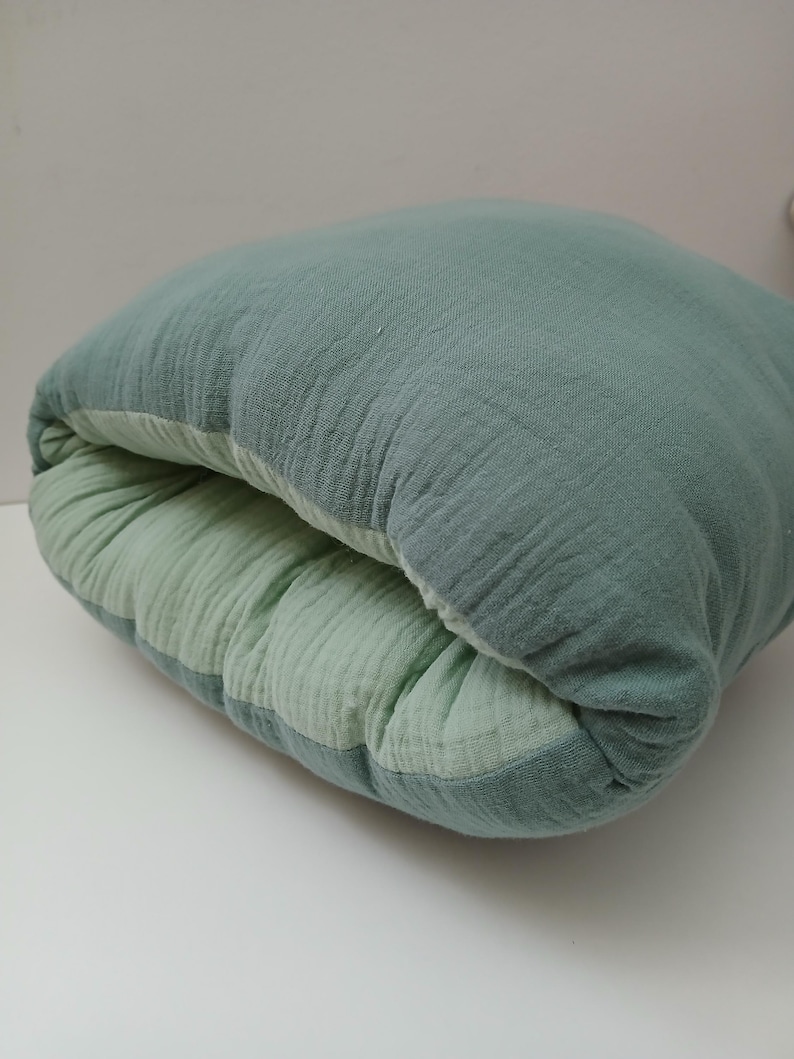 May include: A soft, square-shaped cushion featuring a dual-tone design with a sage green exterior and a lighter mint green interior. The fabric has a visible crinkled, textured finish characteristic of muslin, and the cushion is folded slightly inward.
