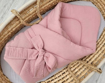 May include: A soft, dusty pink muslin baby swaddle wrap featuring a textured, crinkled fabric finish. The swaddle is designed with a wrap-around style, secured at the front with a large, integrated fabric bow tie, resting inside a woven wicker basket.