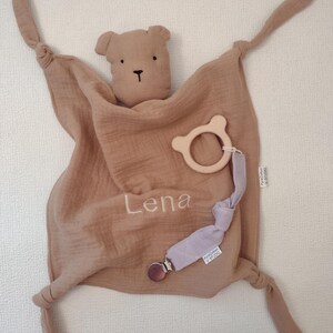 May include: A tan muslin security blanket featuring a bear head, knotted corners, and the name Lena embroidered in white. Attached is a light wooden bear-shaped ring and a lavender fabric strap with a metal clip, both labeled PureCotton STUDIO.