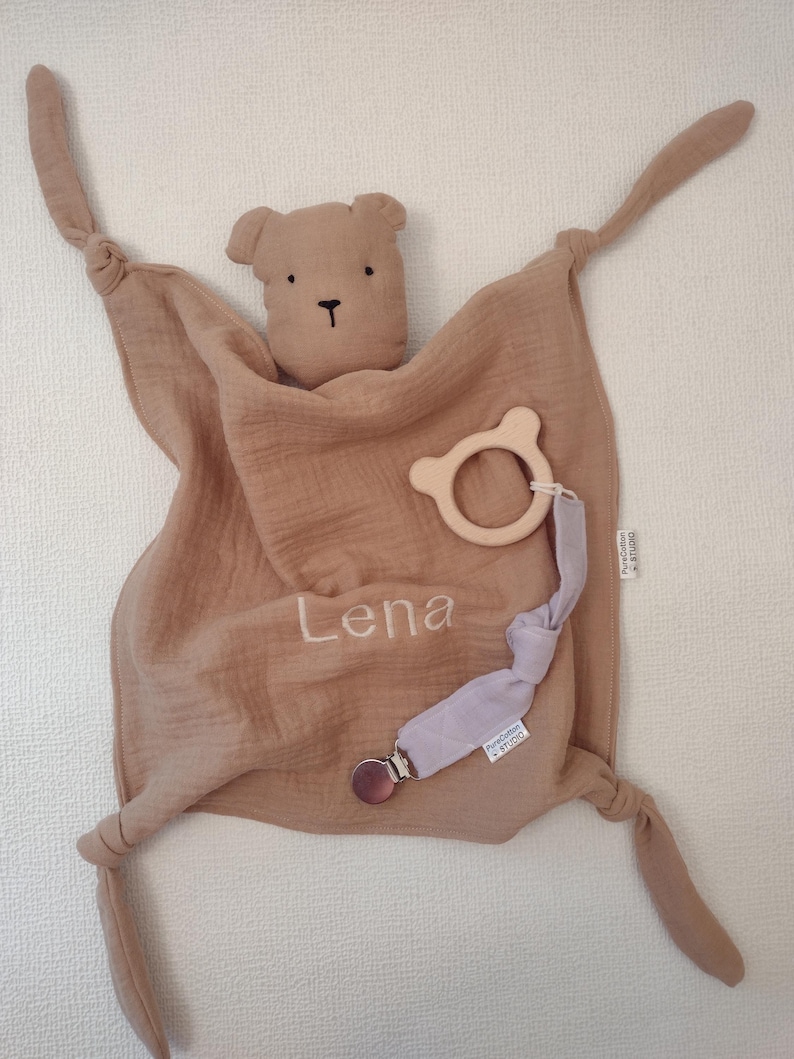 May include: A tan muslin security blanket featuring a bear head, knotted corners, and the name Lena embroidered in white. Attached is a light wooden bear-shaped ring and a lavender fabric strap with a metal clip, both labeled PureCotton STUDIO.