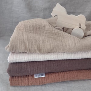May include: A stack of four muslin swaddles in beige, white, brown, and rust colours. The swaddles are folded and stacked on top of each other. A wooden toy horse is on top of the stack. The bottom swaddle has a white label with the text "PureCotton Studio".