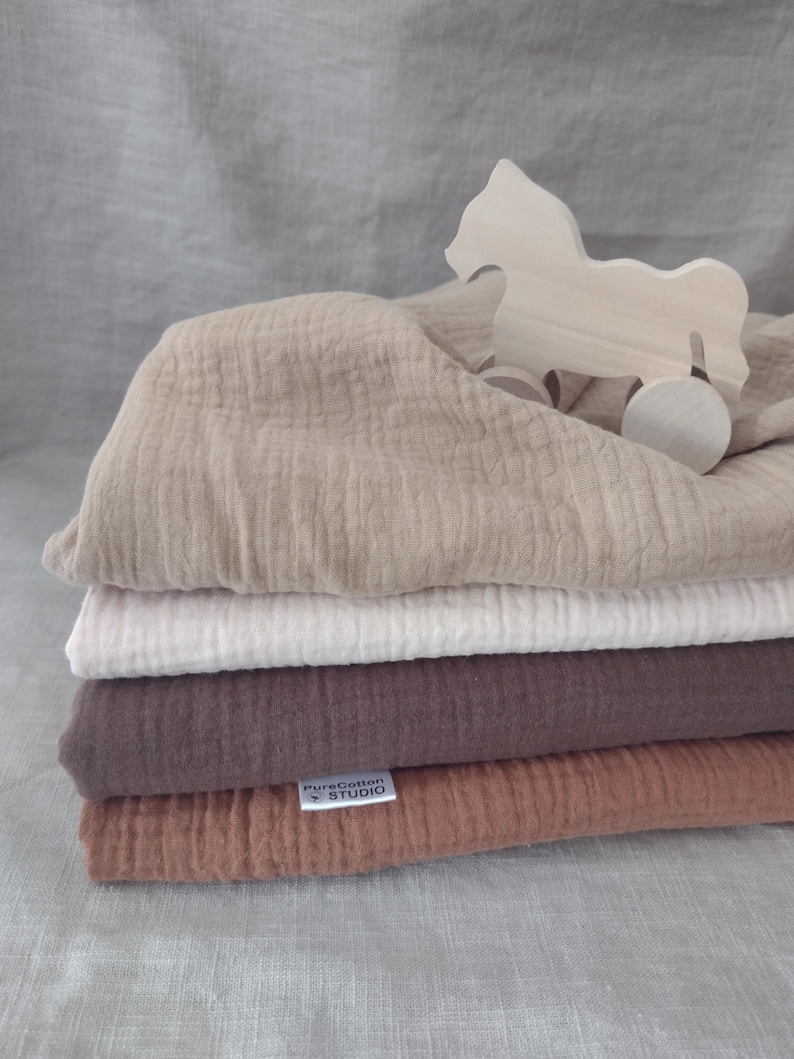 May include: A stack of four muslin swaddles in beige, white, brown, and rust colours. The swaddles are folded and stacked on top of each other. A wooden toy horse is on top of the stack. The bottom swaddle has a white label with the text "PureCotton Studio".