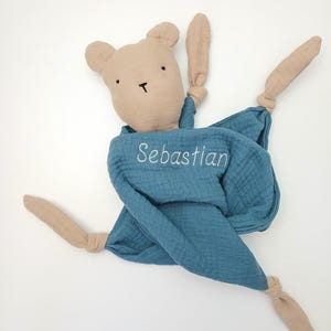 May include: A beige fabric bear head attached to a blue muslin security blanket. The blanket features the embroidered name Sebastian in white thread. The corners of the blue fabric are knotted, creating four long, dangling beige fabric limbs.