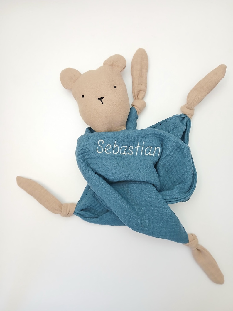 May include: A beige fabric bear head attached to a blue muslin security blanket. The blanket features the embroidered name Sebastian in white thread. The corners of the blue fabric are knotted, creating four long, dangling beige fabric limbs.
