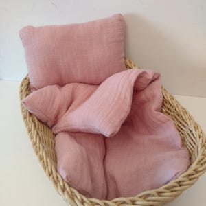 May include: A wicker basket with a pink muslin pillow and blanket. The basket is a light brown color and the fabric is a soft pink.