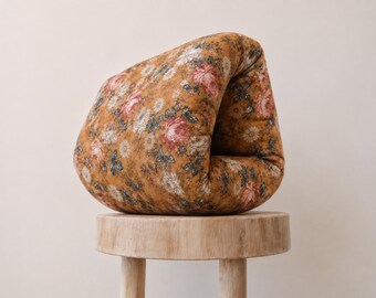 May include: A soft, folded cushion featuring a mustard yellow background with a repeating floral pattern of pink roses, white daisies, and small blue leaves. The fabric has a matte texture and is shaped into a curved, organic, and rounded form.