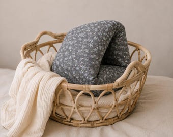 May include: A round woven rattan basket containing a folded dark gray fabric pillow with a small white floral pattern and a cream-colored textured muslin cloth draped over the side of the basket, creating a soft and layered appearance.