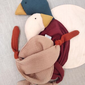 K&ouml;nnte beinhalten: Ein handgefertigtes Pl&uuml;sch-Entenspielzeug mit einem teal-farbenen Kopf, gelbem Schnabel und wei&szlig;em K&ouml;rper. Die Ente hat einen weinroten R&uuml;cken und eine beige, geknotete K&ouml;rperwicklung. Das Spielzeug liegt auf einer hellgrauen Oberfl&auml;che.