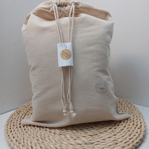 May include: A beige cotton drawstring bag with a rectangular white tag attached to the cords. The tag features a circular sticker with the text "handmade with love". A circular stamp on the lower right side reads "PURE COTTON STUDIO".