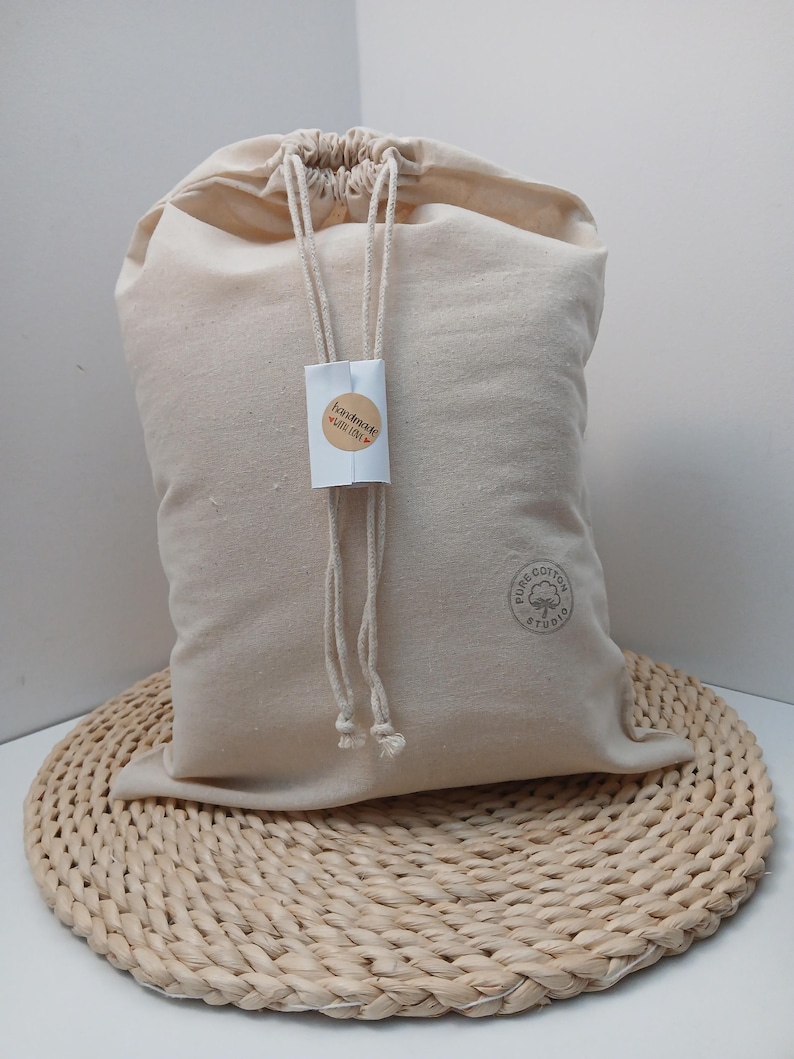 May include: A beige cotton drawstring bag with a rectangular white tag attached to the cords. The tag features a circular sticker with the text "handmade with love". A circular stamp on the lower right side reads "PURE COTTON STUDIO".