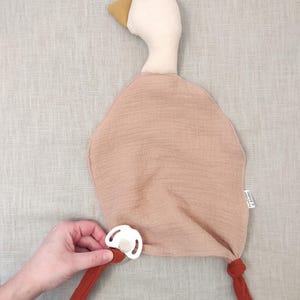 K&ouml;nnte beinhalten: Ein weiches, pl&uuml;schiges Baby-Kuscheltuch in Entenform in Beige- und Rostt&ouml;nen. Die Ente hat einen wei&szlig;en Kopf, einen gelben Schnabel und einen rostfarbenen K&ouml;rper. Ein wei&szlig;er Schnuller ist befestigt. Das Kuscheltuch ist f&uuml;r Babys konzipiert.