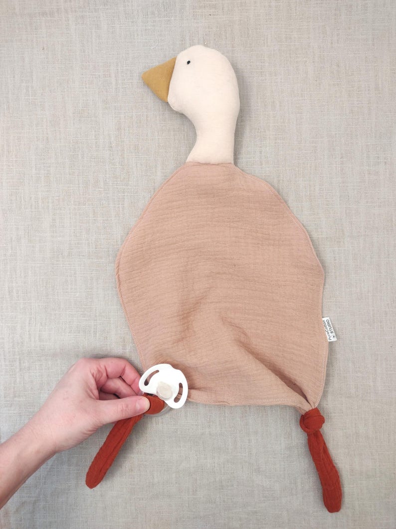 K&ouml;nnte beinhalten: Ein weiches, pl&uuml;schiges Baby-Kuscheltuch in Entenform in Beige- und Rostt&ouml;nen. Die Ente hat einen wei&szlig;en Kopf, einen gelben Schnabel und einen rostfarbenen K&ouml;rper. Ein wei&szlig;er Schnuller ist befestigt. Das Kuscheltuch ist f&uuml;r Babys konzipiert.