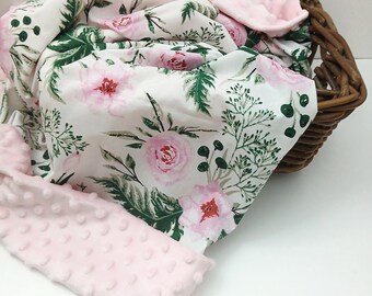 May include: A soft baby blanket featuring a white fabric side with a floral pattern of pink roses and green foliage, paired with a textured light pink minky fabric side with raised dots. The blanket is draped over a brown woven wicker basket.