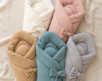 May include: Five baby swaddles made of textured fabric, arranged in a circular pattern. The colors include cream, dusty pink, tan, teal, and light gray. Each swaddle features a soft, padded interior and a decorative fabric bow on the front side.