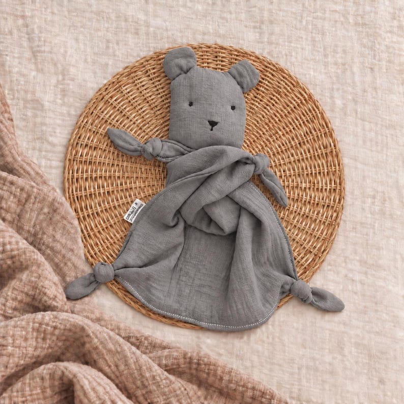 May include: A grey muslin security blanket featuring a plush bear head with embroidered eyes and nose. The fabric has a crinkled texture, with knotted corners on the arms and bottom edges. A small white fabric tag is visible on the side of the bear.