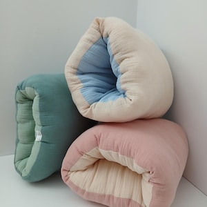 May include: Three soft, rectangular, folded fabric cushions made of layered muslin material. One cushion is sage green with a lighter green interior, one is cream with a light blue interior, and one is dusty rose with a cream interior.
