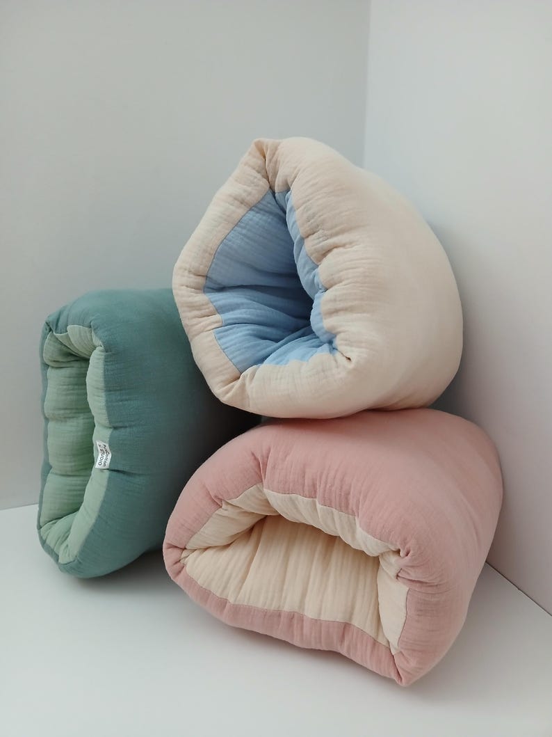 May include: Three soft, rectangular, folded fabric cushions made of layered muslin material. One cushion is sage green with a lighter green interior, one is cream with a light blue interior, and one is dusty rose with a cream interior.