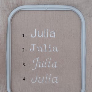 May include: A close-up of four embroidered examples of the name "Julia" in white thread on a tan fabric. The name is displayed in different fonts. The fabric is held in a white embroidery hoop.