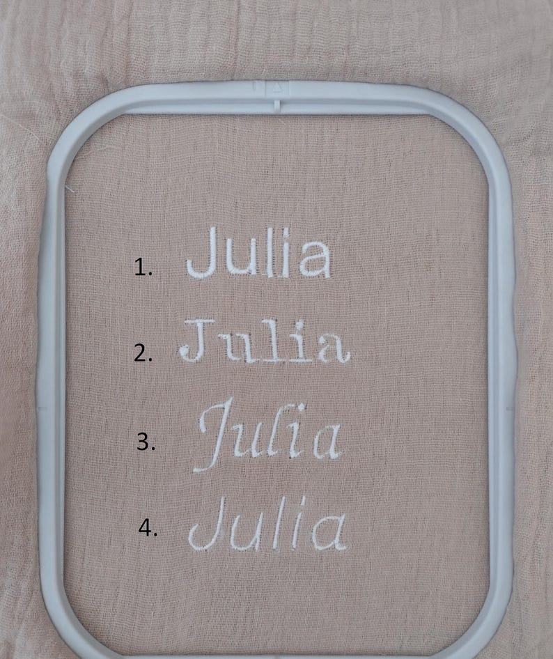 May include: A close-up of four embroidered examples of the name "Julia" in white thread on a tan fabric. The name is displayed in different fonts. The fabric is held in a white embroidery hoop.