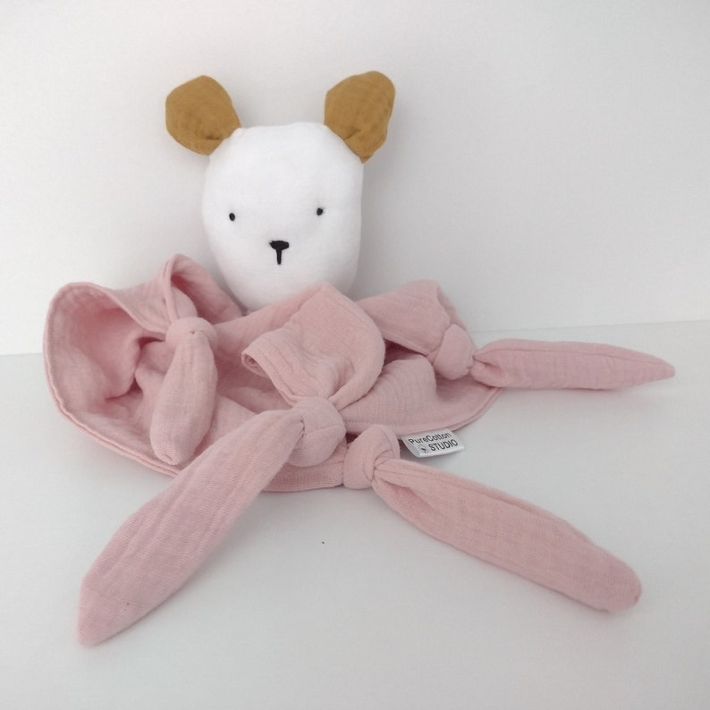May include: A soft toy featuring a white head with mustard yellow ears and a pink muslin fabric body. The pink fabric has four knotted corners and a small white tag that reads PureCotton STUDIO. The toy has simple black embroidered facial features.