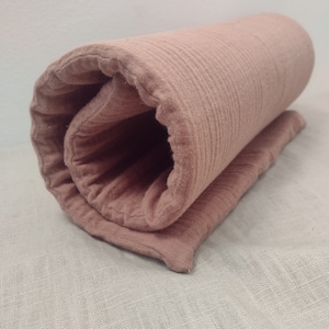 May include: A rolled up blush pink muslin swaddle blanket with a textured surface.