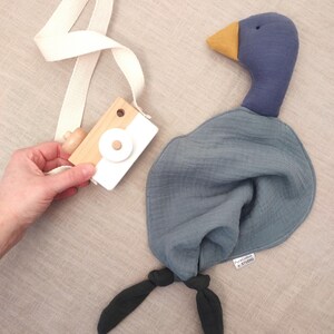 K&ouml;nnte beinhalten: Eine Holzspielzeugkamera mit wei&szlig;em Riemen wird neben einer blauen Stoffente gehalten. Die Ente hat einen gelben Schnabel, einen blauen Kopf und K&ouml;rper sowie dunkelgr&uuml;ne F&uuml;&szlig;e. Die Ente hat ein Etikett mit der Aufschrift "PureCotton STUDIO".