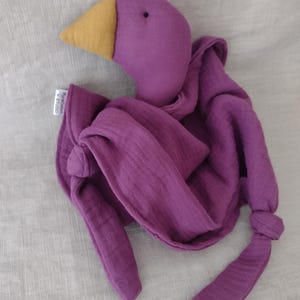 May include: A purple plush bird toy with a yellow beak and a soft, draped body. The bird's body is made of a textured fabric, and the design includes long, knotted appendages. A small tag is attached to the bird.