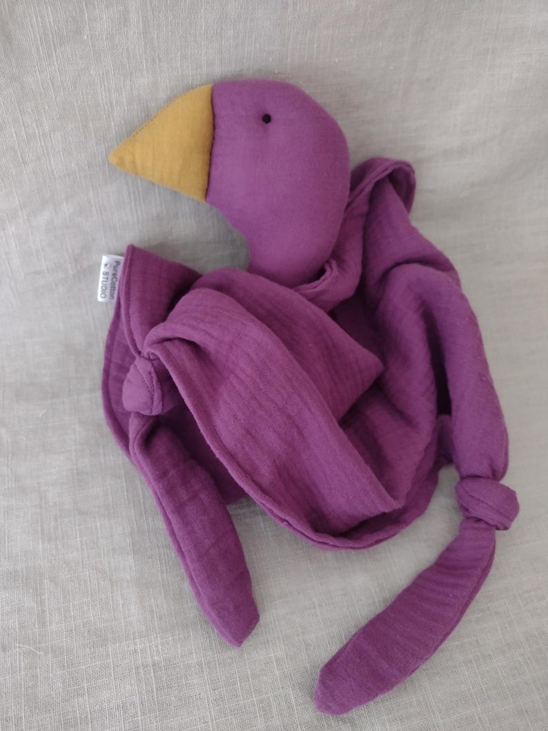 May include: A purple plush bird toy with a yellow beak and a soft, draped body. The bird's body is made of a textured fabric, and the design includes long, knotted appendages. A small tag is attached to the bird.