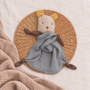 May include: A baby security blanket featuring a beige bear head with mustard yellow ears and a dark brown knotted arm on each side. The body of the blanket is made of a soft, crinkled grey fabric that drapes in folds, creating a textured appearance.