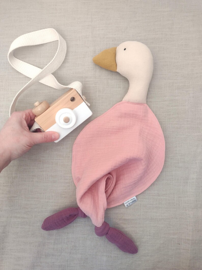 K&ouml;nnte beinhalten: Ein Stofftier in Form einer Gans mit rosa K&ouml;rper und lila F&uuml;&szlig;en, neben einer Holzspielzeugkamera mit wei&szlig;em Riemen. Die Gans hat einen cremefarbenen Kopf und einen gelben Schnabel. Eine Hand h&auml;lt die Kamera.