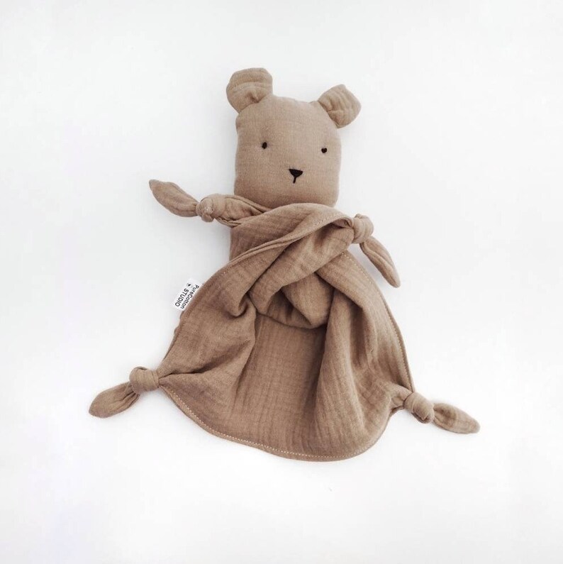 May include: A light brown muslin security blanket featuring a plush bear head with embroidered black eyes and nose. The fabric has a crinkled texture with four knotted corners and a small white fabric tag that reads PureCotton STUDIO.
