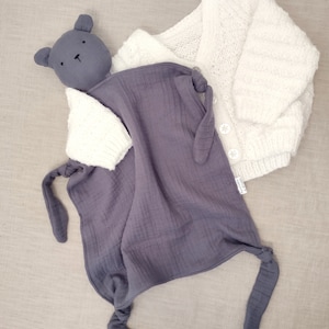 May include: A dark gray muslin security blanket featuring a bear head and knotted corners, paired with a white long-sleeve knitted cardigan that has a textured pattern and three white buttons down the front closure.