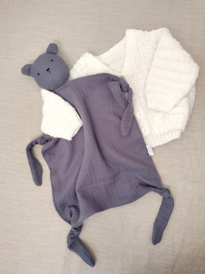 May include: A dark gray muslin security blanket featuring a bear head and knotted corners, paired with a white long-sleeve knitted cardigan that has a textured pattern and three white buttons down the front closure.