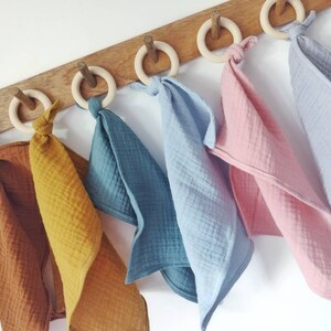 May include: Six muslin baby teethers hanging on a wooden rack. The teethers are in various colors, including brown, mustard yellow, teal, light blue, and pink. Each teether is attached to a wooden ring.