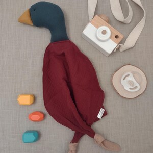 K&ouml;nnte beinhalten: Ein Stofftier-Ente mit einem teal-farbenen Kopf und einem weinroten K&ouml;rper, zusammen mit einer Spielzeugkamera aus Holz, einem Schnuller und drei bunten geometrischen Bl&ouml;cken. Die Ente hat einen gelben Schnabel und beige F&uuml;&szlig;e. Die Kamera hat eine wei&szlig;e Vorderseite und einen beigen Riemen.