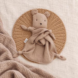 May include: A beige muslin security blanket featuring a stuffed bear head with embroidered eyes and nose. The soft fabric body has knotted corners and a small white brand tag attached to the side, creating a simple and tactile comfort object.