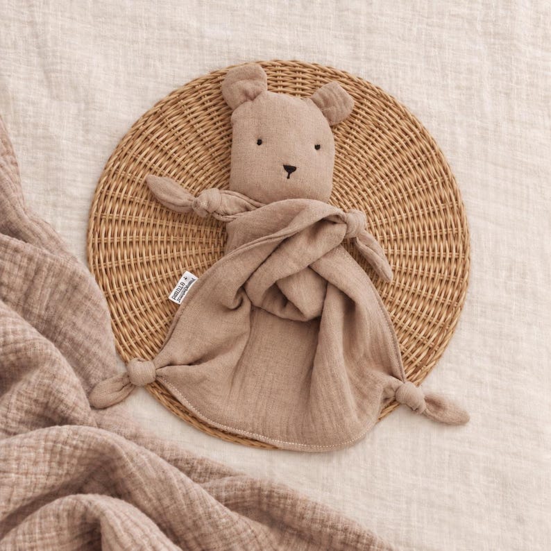 May include: A beige muslin security blanket featuring a stuffed bear head with embroidered eyes and nose. The soft fabric body has knotted corners and a small white brand tag attached to the side, creating a simple and tactile comfort object.