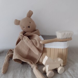 May include: A tan muslin security blanket with a stuffed animal head, a light wood hairbrush with white bristles, a small wooden toy horse on wheels, and a rectangular bamboo storage basket with a white fabric liner and a small white tag.