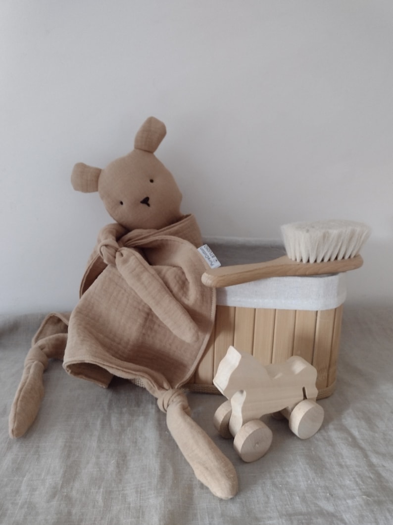 May include: A tan muslin security blanket with a stuffed animal head, a light wood hairbrush with white bristles, a small wooden toy horse on wheels, and a rectangular bamboo storage basket with a white fabric liner and a small white tag.