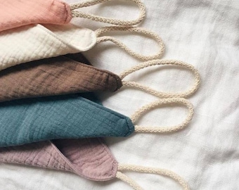 May include: A stack of five triangular fabric cloths made of textured muslin material in shades of peach, cream, brown, teal, and mauve. Each cloth features a stitched edge and a beige braided rope loop attached to one corner for hanging purposes.