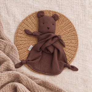 May include: A dark brown muslin security blanket featuring a plush bear head with embroidered black eyes and nose. The soft fabric body has knotted corners and a small white brand tag on the side, creating a simple, tactile comfort object.