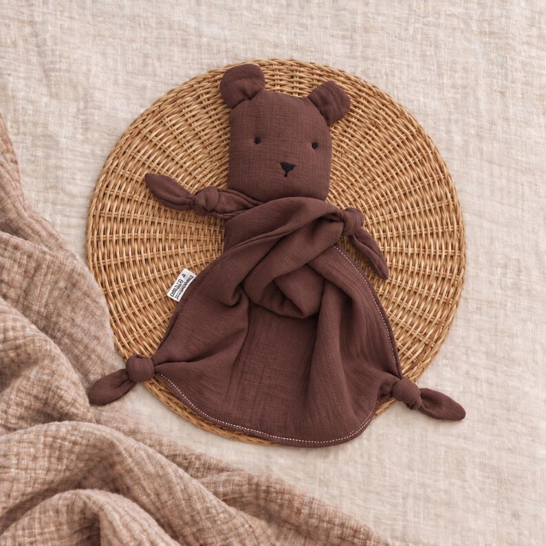 May include: A dark brown muslin security blanket featuring a plush bear head with embroidered black eyes and nose. The soft fabric body has knotted corners and a small white brand tag on the side, creating a simple, tactile comfort object.