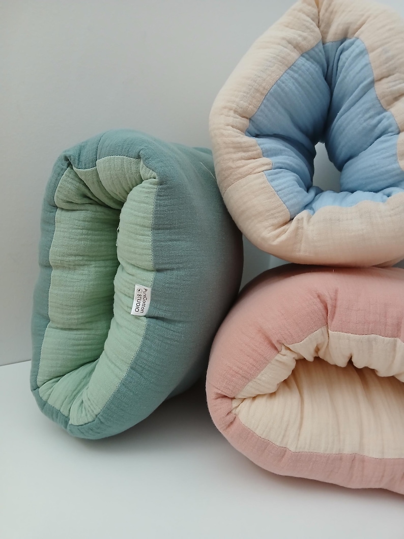 May include: Three soft, tubular cushions made of crinkled fabric. One is sage green with a light green interior, one is beige with a light blue interior, and one is dusty pink with a cream interior. A white tag reads PureCotton STUDIO.