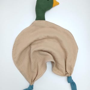 May include: A plush duck-shaped baby comforter. The duck has a green head, a yellow beak, and a tan body. The comforter has two teal-colored knotted legs. The soft toy is designed for infants and toddlers.