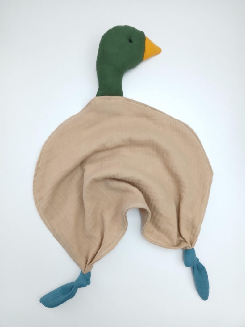 May include: A plush duck-shaped baby comforter. The duck has a green head, a yellow beak, and a tan body. The comforter has two teal-colored knotted legs. The soft toy is designed for infants and toddlers.