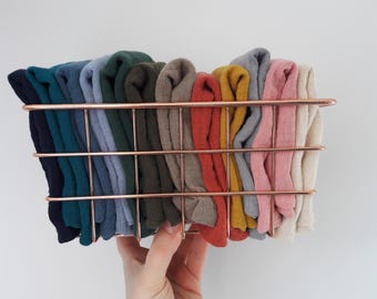 May include: A rectangular rose gold wire basket filled with a collection of folded fabric swatches in various colors including navy, teal, blue, forest green, olive, taupe, rust, mustard, grey, dusty pink, and cream, arranged in a row.